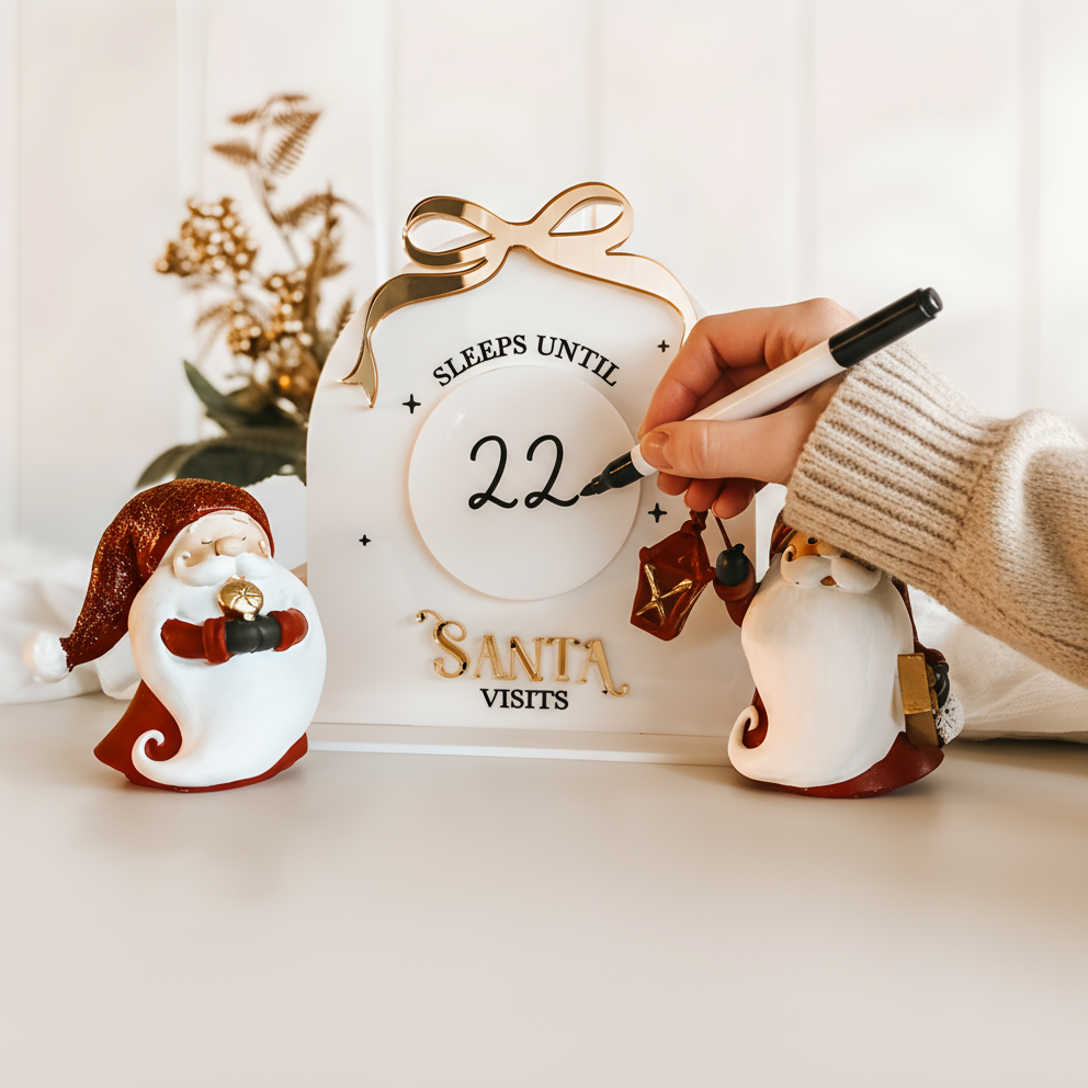 Decorative clock with 'Sleeps Until Santa Visits' text, surrounded by festive decor on a light surface.