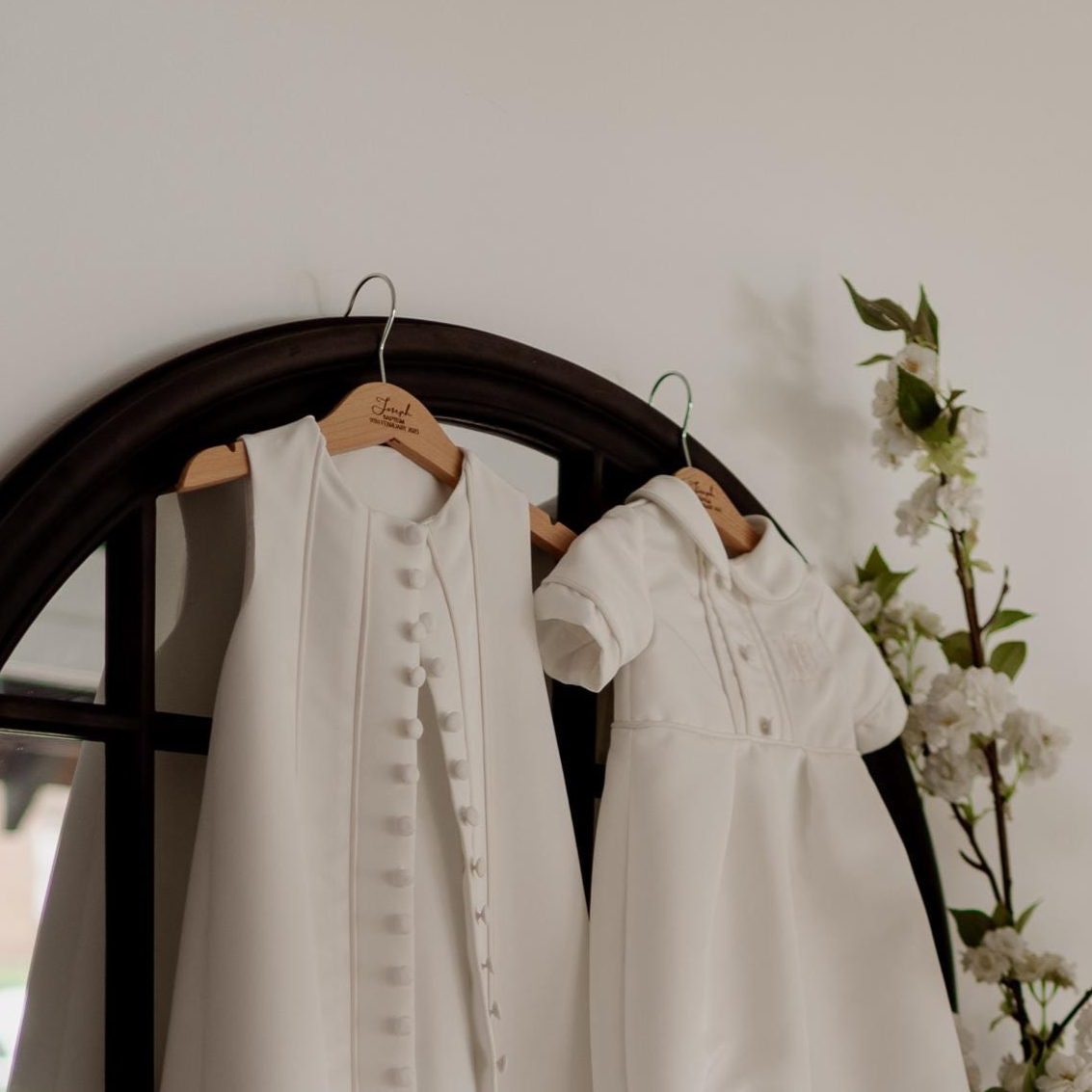 White baby christening outfit on custom engraved coat hangers with acrylic cross in front of a round mirror with floral decorations