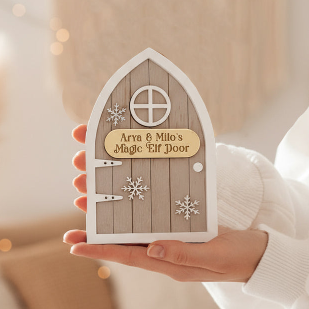 Woman holding a Handcrafted personalised Magic Elf Door made from wood and acrylic, featuring a gold nameplate, 3D snowflake accents, and arched brick detailing. A beautiful Christmas decoration designed to welcome Santa’s little helper into your home. Perfect for family traditions, children’s imagination, and festive storytelling throughout December.