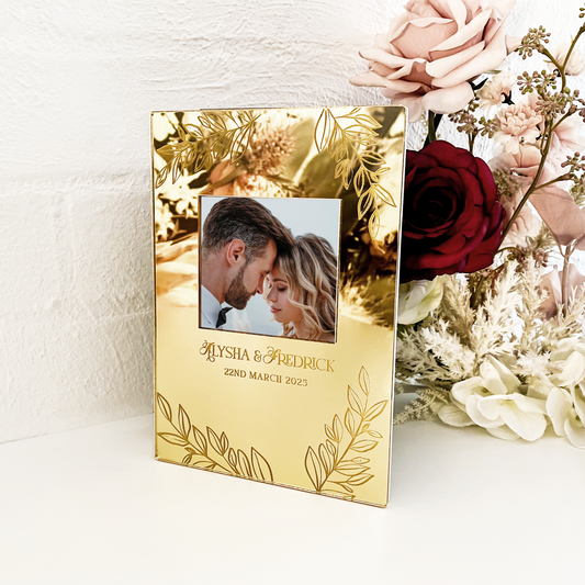 Gold photo frame with a couple's photo and personalized text, next to a bouquet of flowers on a white surface.