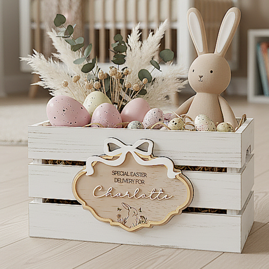 A trending coquette-style Easter crate sign made of 3mm plywood with a 3D white acrylic name and a matching 3D white bow. The plaque has a luxury gold metallic border and is attached to a wooden crate filled with pastel eggs. The "Special Easter Delivery" engraving and dainty bunny motif create a whimsical, high-end aesthetic for a personalized Easter morning surprise.