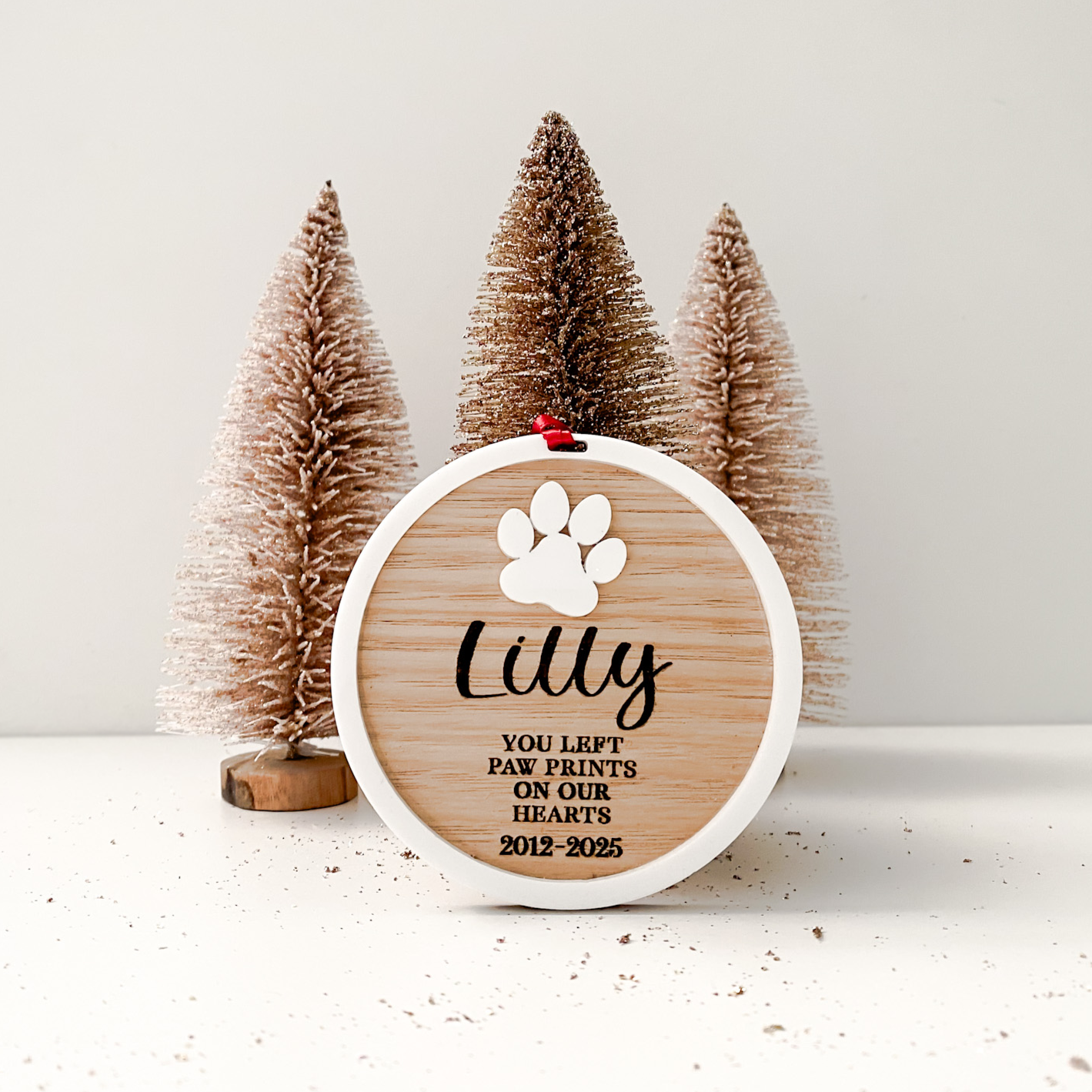 Decorative plaque with paw print design and text in front of small Christmas trees on a white background
