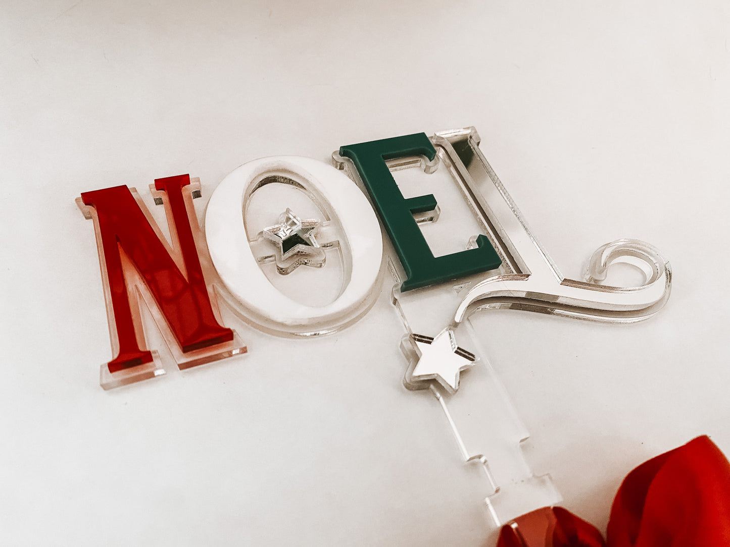 Modern acrylic Noel tree topper handcrafted by Bell & Beau Design Co, featuring layered red, white, and green acrylic letters with a bright red bow for easy tree attachment. A stylish holiday statement piece designed to complement both classic and contemporary Christmas themes.