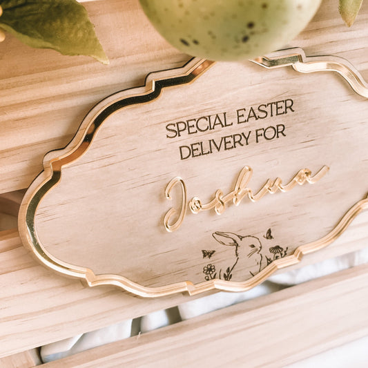 A custom wooden Easter basket plaque featuring the name "Joshua" with a metallic gold border. This "no bow" version is styled on a wooden treat crate in a modern living room alongside a plush bunny and a Peter Rabbit book. The 20cm sign is a perfect example of a timeless Easter tradition, designed to be used year after year as a durable, high-quality family keepsake.