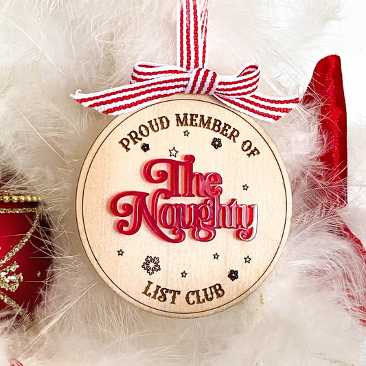 A close-up of the Naughty List Ornament, a wooden Christmas decoration with red 3D acrylic text, hanging on a frosty Christmas tree branch