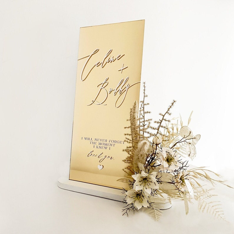 Vertical gold mirror acrylic wedding welcome sign featuring custom names, a quote, and a floral arrangement.