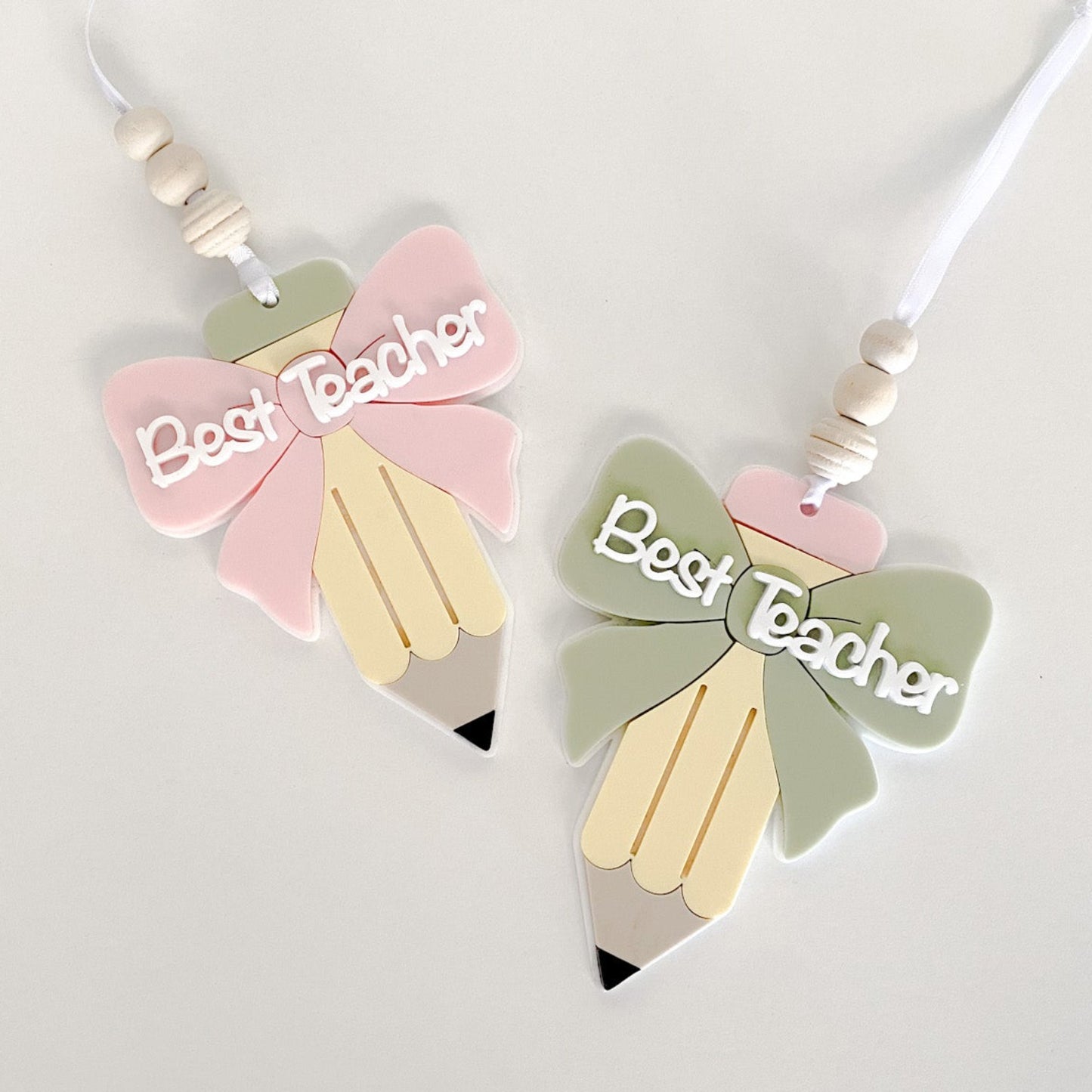 Two layered acrylic teacher appreciation ornaments in a pencil shape with a pink bow and a sage green bow.